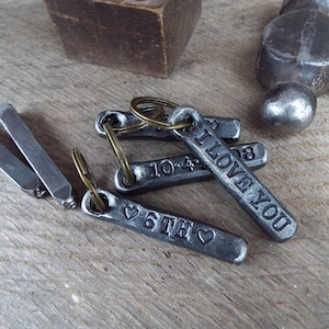 May include: Several metal keychains with stamped text and dates, including "I LOVE YOU" and "6TH". The keychains are silver-colored and attached to brass-colored key rings. The background is a wooden surface with tools.