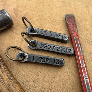 May include: Three metal keychains with stamped text: "1 TH 5:16-18", "PROV 27:17", and "1 COR. 13". A hammer and a red chisel are also visible. The items are on a wooden surface.