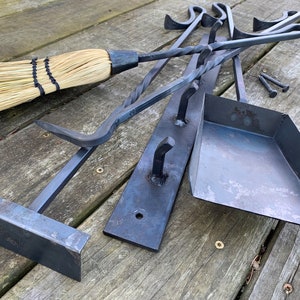 May include: A set of black metal fireplace tools with a matching shovel and a broom. The tools are hanging on a black metal rack with hooks. The tools are on a wooden surface.