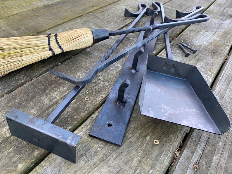 May include: A set of black metal fireplace tools with a matching shovel and a broom. The tools are hanging on a black metal rack with hooks. The tools are on a wooden surface.