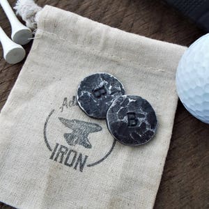 May include: Two small, round, metal golf ball markers with the letter "B" stamped on them. The markers are in a small, white canvas bag with the words "Artisan Iron" printed on it.