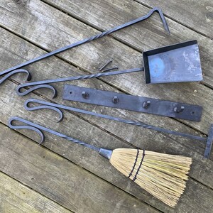 May include: A set of fireplace tools with a black metal finish, including a poker, shovel, tongs, and a brush. The tools are hanging on a black metal rack with hooks. The tools are on a wooden surface.