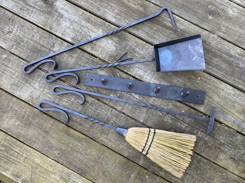 May include: A set of fireplace tools with a black metal finish, including a poker, shovel, tongs, and a brush. The tools are hanging on a black metal rack with hooks. The tools are on a wooden surface.