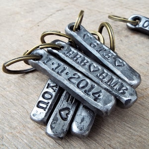 May include: A collection of personalized metal keychains with stamped text and dates. The keychains are rectangular with rounded edges and attached to bronze-colored key rings. The text includes names, initials, dates, and heart symbols.