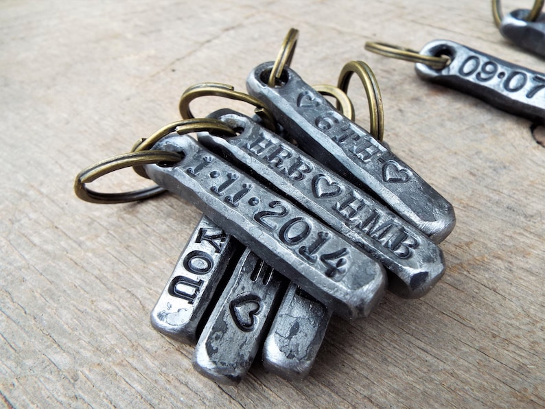 May include: A collection of personalized metal keychains with stamped text and dates. The keychains are rectangular with rounded edges and attached to bronze-colored key rings. The text includes names, initials, dates, and heart symbols.
