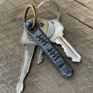 May include: A set of keys with a hand-forged, dark gray metal keychain. The keychain has a series of etched lines and is attached to a gold-colored ring. The keys are silver and gold-toned, resting on a wooden surface.