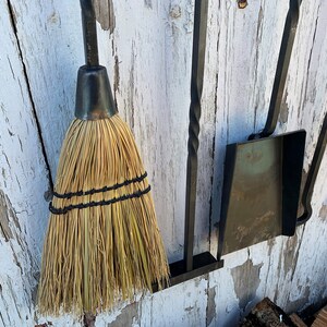 May include: A set of fireplace tools hanging on a white wooden wall. The tools include a broom, a shovel, and a poker. The tools are made of black metal and have a rustic look.