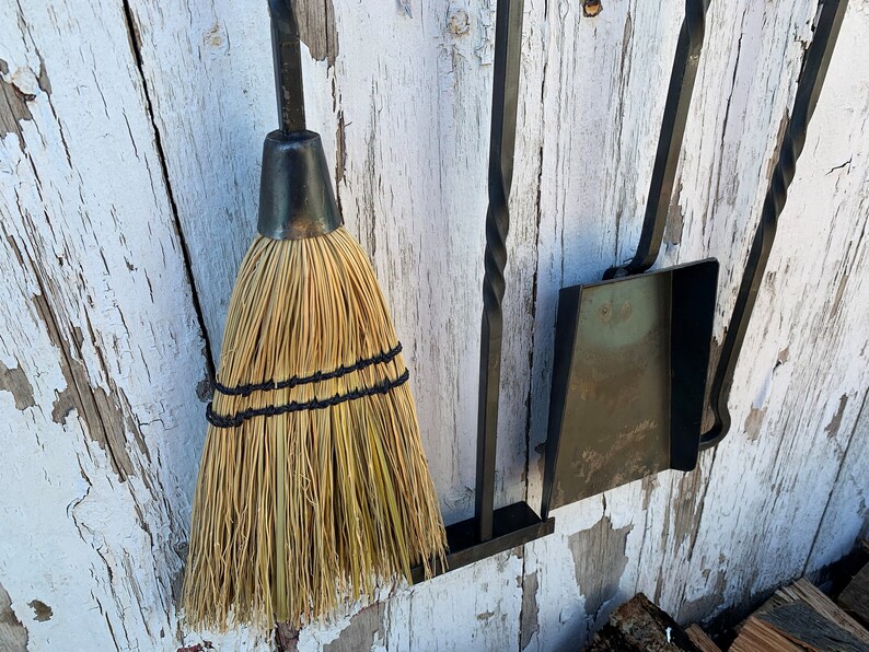 May include: A set of fireplace tools hanging on a white wooden wall. The tools include a broom, a shovel, and a poker. The tools are made of black metal and have a rustic look.