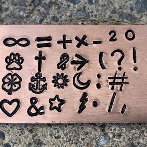 May include: A copper-colored metal plate with various symbols and characters stamped into it. Symbols include an infinity sign, paw print, cross, clover, anchor, heart, ampersand, star, lightning bolt, and mathematical symbols.