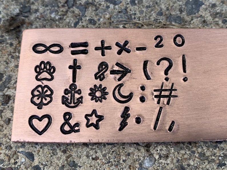 May include: A copper-colored metal plate with various symbols and characters stamped into it. Symbols include an infinity sign, paw print, cross, clover, anchor, heart, ampersand, star, lightning bolt, and mathematical symbols.