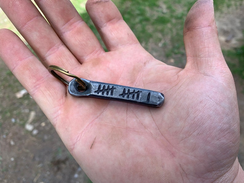 May include: A hand-forged metal keychain with a brass ring. The rectangular tag features etched tally marks, indicating a count of 11. The metal has a dark, textured finish, and the keychain is held in an open palm.