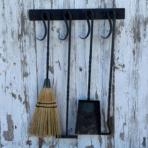 May include: A set of four black metal fireplace tools hanging on a white wooden wall. The tools include a brush, a shovel, a poker, and a tongs. The tools are all attached to a black metal bar with hooks.