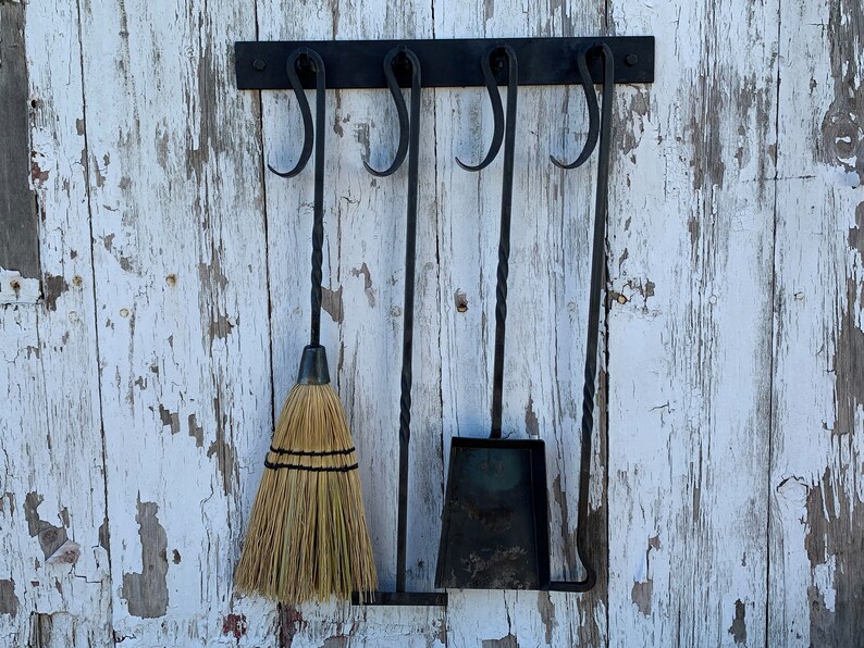 May include: A set of four black metal fireplace tools hanging on a white wooden wall. The tools include a brush, a shovel, a poker, and a tongs. The tools are all attached to a black metal bar with hooks.