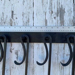 May include: Black metal wall-mounted hook rack with four hooks. The rack is mounted on a white wooden surface. A ruler is placed above the rack, showing measurements in inches and millimeters.