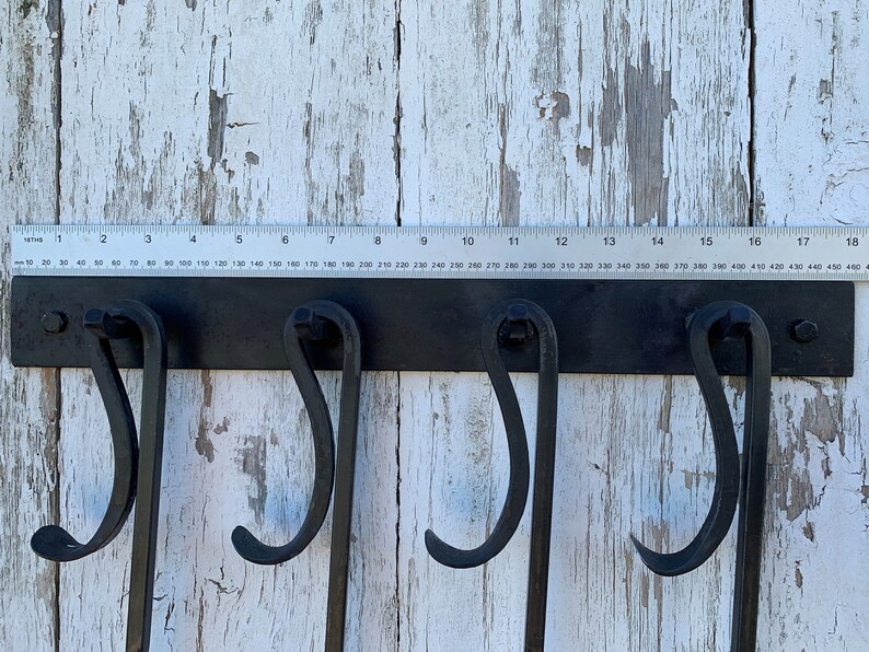May include: Black metal wall-mounted hook rack with four hooks. The rack is mounted on a white wooden surface. A ruler is placed above the rack, showing measurements in inches and millimeters.
