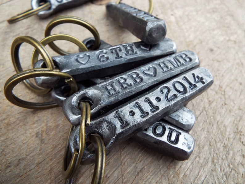 May include: Close-up of several metal keychains with engraved text and dates. The keychains are attached to brass-colored key rings. The text includes initials, dates, and a heart symbol. The keychains are made of a dark gray metal.