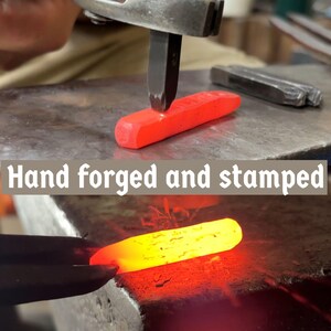 May include: A blacksmith forges a piece of red-hot metal with a hammer and stamp. The text "Hand forged and stamped" is displayed. The image shows the process of metalworking, highlighting the heat and force involved in the craft.