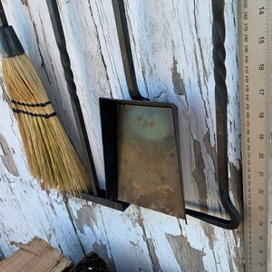 May include: A black metal fireplace tool set with a broom, shovel, and poker. The tools are mounted on a wall with a wooden ruler next to them.