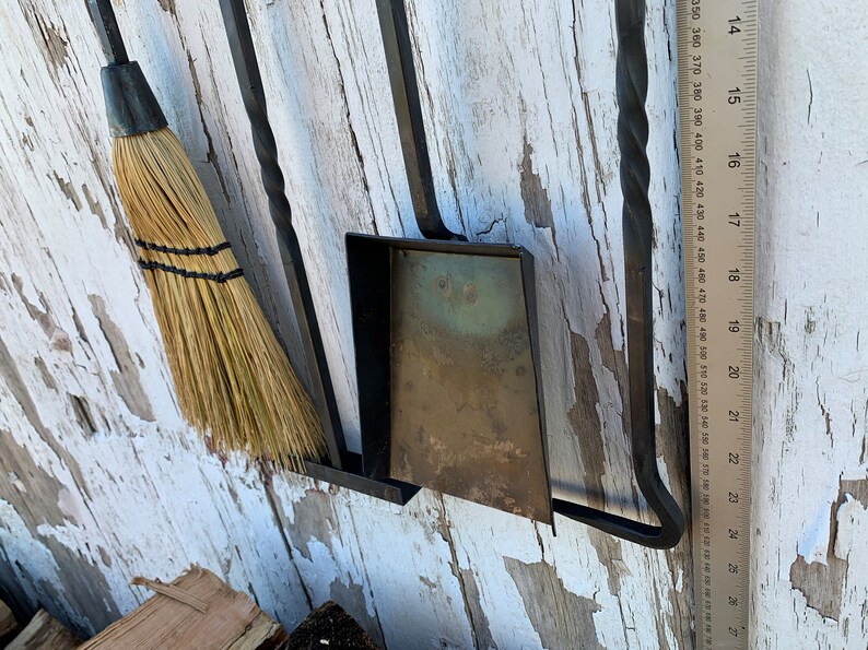 May include: A black metal fireplace tool set with a broom, shovel, and poker. The tools are mounted on a wall with a wooden ruler next to them.