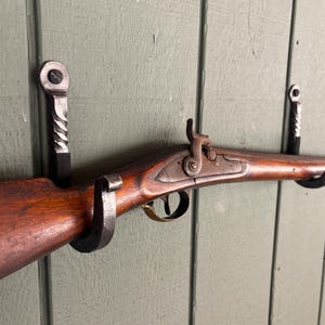 May include: A wooden rifle with metal accents is displayed on a wall mounted gun rack. The rack is made of twisted metal and has a dark gray finish.