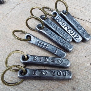May include: A collection of personalized metal keychains with bronze-colored key rings. Each rectangular keychain is engraved with dates, initials, and the phrase "I ❤️ YOU". The keychains are arranged on a wooden surface.