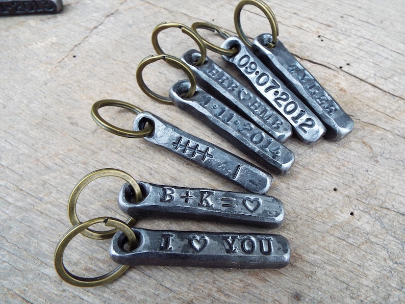 May include: A collection of personalized metal keychains with bronze-colored key rings. Each rectangular keychain is engraved with dates, initials, and the phrase "I ❤️ YOU". The keychains are arranged on a wooden surface.