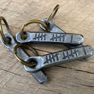 May include: Three metal keychains with bronze-colored key rings. Each keychain is a rectangular shape with a series of etched tally marks and a single vertical line. The keychains are made of a dark, textured metal.