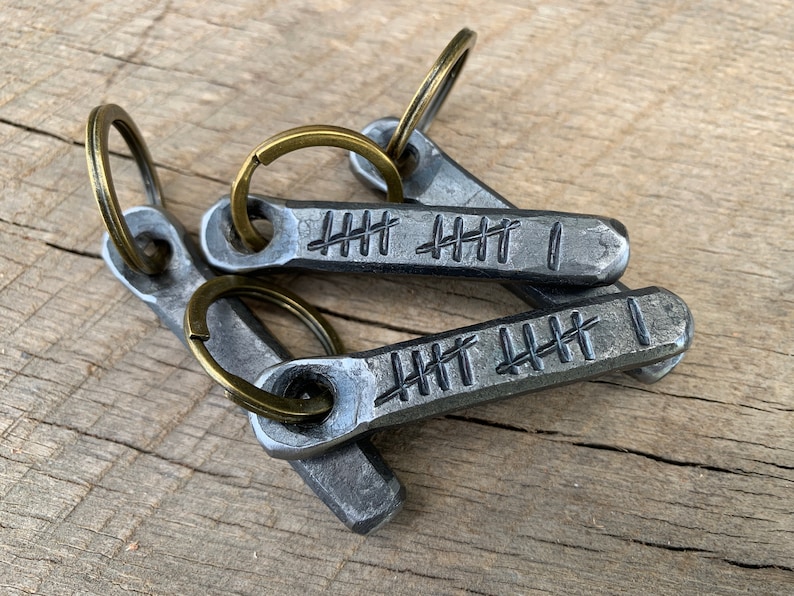 May include: Three metal keychains with bronze-colored key rings. Each keychain is a rectangular shape with a series of etched tally marks and a single vertical line. The keychains are made of a dark, textured metal.