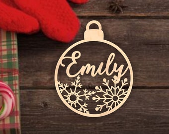 May include: A light-colored wooden Christmas ornament shaped like a round bauble, featuring the name Emily in cursive script and two intricate snowflake cutouts at the bottom, displayed against a dark brown rustic wooden surface.