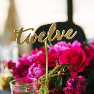 以下が含まれることがあります： 「twelve」と書いた、筆記体で書かれた金のテーブルナンバーカード。カードはピンクと赤のバラのブーケに置かれています。