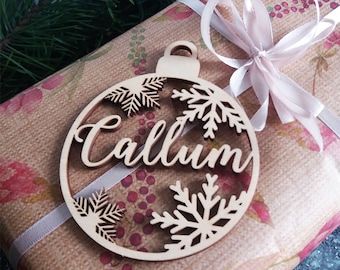 May include: A wooden Christmas ornament shaped like a bauble with the name Callum cut out in the center. The ornament is decorated with snowflakes and is sitting on top of a brown gift box with a pink ribbon.