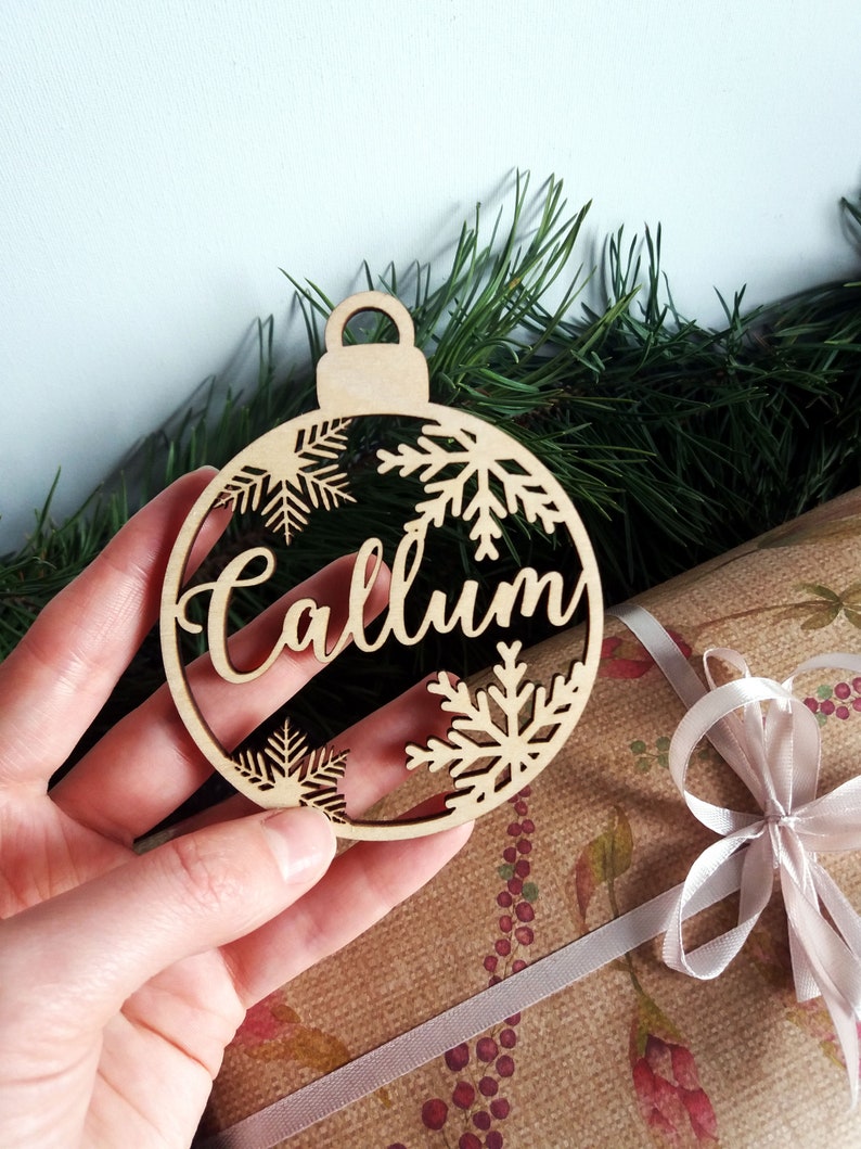 May include: A wooden Christmas ornament shaped like a bauble with the name "Callum" cut out in the center. The ornament is decorated with snowflake cutouts.