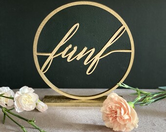 May include: A gold-colored wooden sign with the word "funf" in a cursive font, set within a black circle. The sign is on a gold base. Soft pink flowers are arranged around the sign on a gray surface.