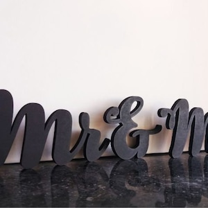 May include: Black wooden cutout letters spelling "Mr. & Mrs." in a cursive font. The letters are arranged in a line on a black surface.