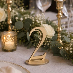 May include: A gold-colored table number sign featuring the numeral 2 in a stylized, modern font. The sign is constructed with a flat, circular base that supports the upright, cut-out number, providing a stable display for event table settings.