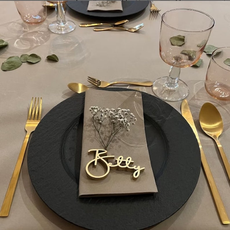 Puede incluir: Un plato de pizarra negro con una tarjeta de lugar dorada que dice "Betty" sobre una servilleta marr&oacute;n con flores blancas de aliento de beb&eacute;. El plato est&aacute; colocado sobre un mantel beige con cubiertos dorados y un vaso rosa.