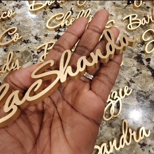 Puede incluir: Una mano sosteniendo un letrero de madera dorado con el nombre "Shanda" escrito en cursiva. El letrero est&aacute; rodeado de otros letreros de madera dorados con nombres escritos en ellos.