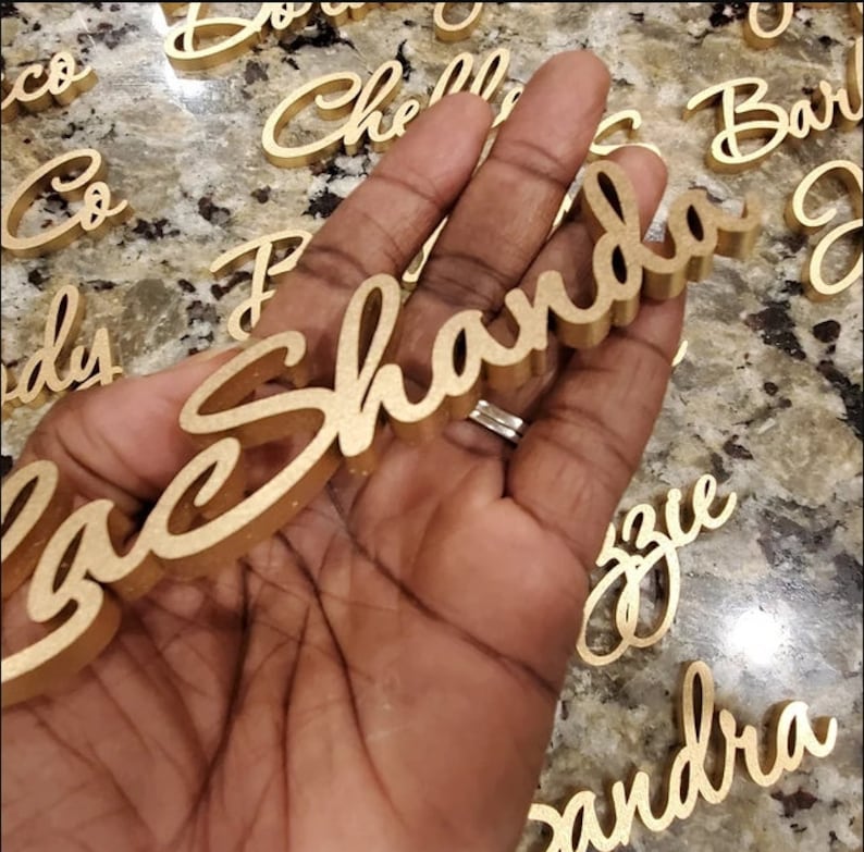 Puede incluir: Una mano sosteniendo un letrero de madera dorado con el nombre "Shanda" escrito en cursiva. El letrero est&aacute; rodeado de otros letreros de madera dorados con nombres escritos en ellos.