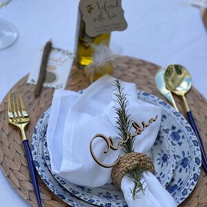 Puede incluir: Una servilleta blanca con una tarjeta de nombre de madera que dice "Claudia" y un anillo de servilleta tejido. La servilleta est&aacute; colocada sobre un plato con estampado azul y blanco con un plato de servicio tejido. Una peque&ntilde;a botella de aceite de oliva con una etiqueta que dice "Infundido con amor" est&aacute; en el fondo.