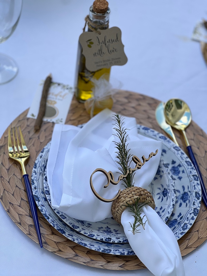 Puede incluir: Una servilleta blanca con una tarjeta de nombre de madera que dice "Claudia" y un anillo de servilleta tejido. La servilleta est&aacute; colocada sobre un plato con estampado azul y blanco con un plato de servicio tejido. Una peque&ntilde;a botella de aceite de oliva con una etiqueta que dice "Infundido con amor" est&aacute; en el fondo.