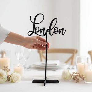 Könnte beinhalten: Ein schwarzes Tischschild mit dem Wort "London" in Schreibschrift. Das Schild wird von einer Hand gehalten. Das Schild steht auf einem Tisch mit Kerzen, Blumen und Glaswaren.