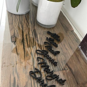 Puede incluir: Tarjetas de lugar de madera negra sobre una mesa de madera con veta de madera clara. Los nombres son "Lillian", "Lisa", "Grandma", "Margaret" y "David".