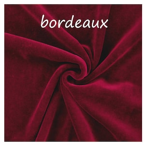 Op de afbeelding: Een diepbordeauxrode fluwelen stof met een zachte, pluche textuur en natuurlijke plooien en draperingen. Het woord "bordeaux" is in witte, kleine, schreefloze letters gedrukt nabij de bovenkant van het stoffen oppervlak.