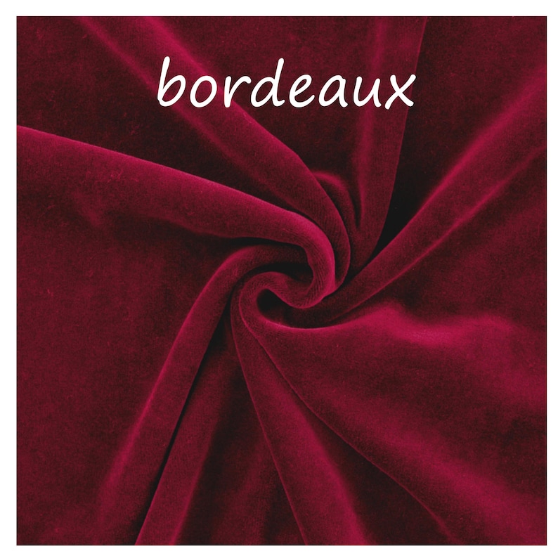 Op de afbeelding: Een diepbordeauxrode fluwelen stof met een zachte, pluche textuur en natuurlijke plooien en draperingen. Het woord "bordeaux" is in witte, kleine, schreefloze letters gedrukt nabij de bovenkant van het stoffen oppervlak.