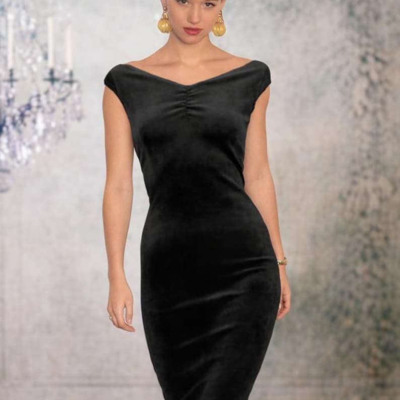 Op de afbeelding: Een zwarte fluwelen jurk met een brede, off-shoulder halslijn en een subtiel geplooid detail in het midden aan de voorzijde. Het kledingstuk is getailleerd bij de romp en taille en loopt uit in een rechte, knielange rok met korte mouwen.