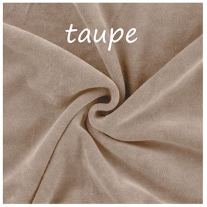 Op de afbeelding: Een stuk taupekleurige stof met een zachte, fluweelachtige textuur. Het materiaal is in het midden samengevouwen, waardoor diepe plooien en schaduwen ontstaan. Het woord taupe is in witte, kleine, schreefloze letters bovenaan gedrukt.