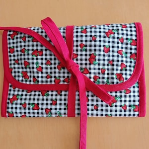 Puede incluir: Un estuche enrollable de tela vichy roja y blanca con un lazo rojo y estampado de fresas.