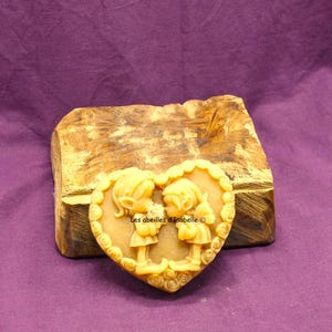 Puede incluir: Una vela de cera de abeja en forma de corazón con un diseño en relieve de dos figuras que se toman de la mano. La vela es de color amarillo claro y tiene un diseño en relieve de rosas alrededor del borde. El texto "Les abeilles d'Isabelle ©" está impreso en la vela.