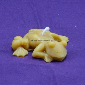 May include: A yellow beeswax frog candle with a white wick. The candle is lying on its side on a purple surface. The text "Les abeilles d'Isabelle ©" is visible on the candle.