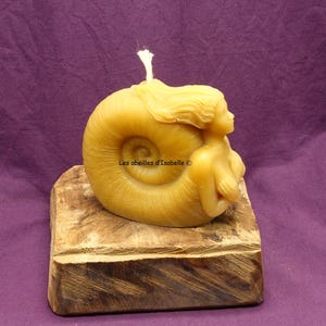 Puede incluir: Una vela de cera de abeja con forma de sirena sentada dentro de una concha. La vela está sobre una base de madera. La vela es de color amarillo claro. El texto "Les abeilles d'Isabelle ©" está en la vela.